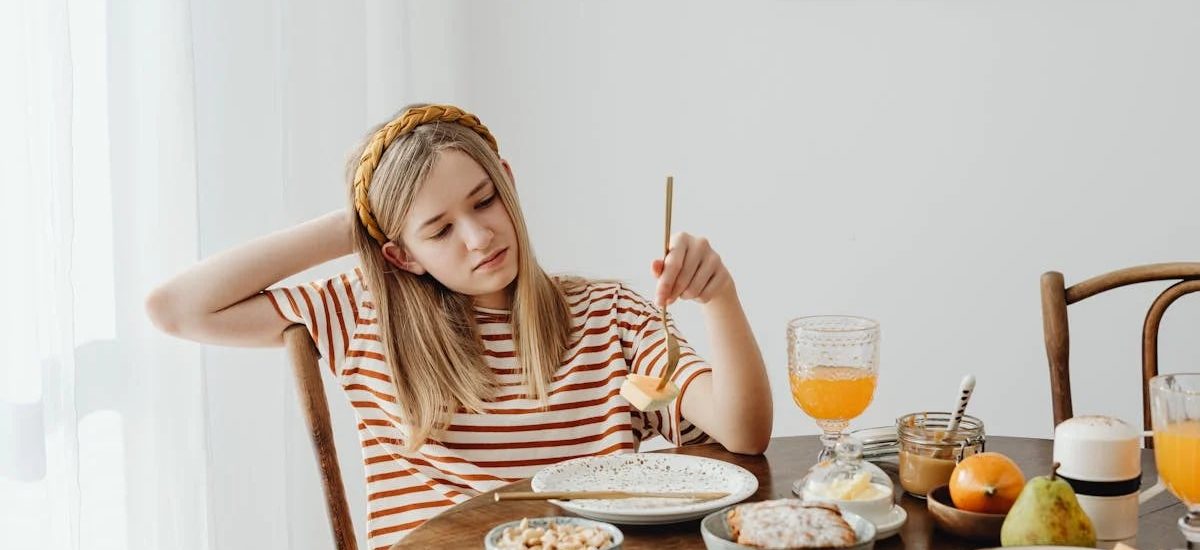 Teen girl in striped shirt at breakfast table with fruits, bread, and juice. Casual morning setting.